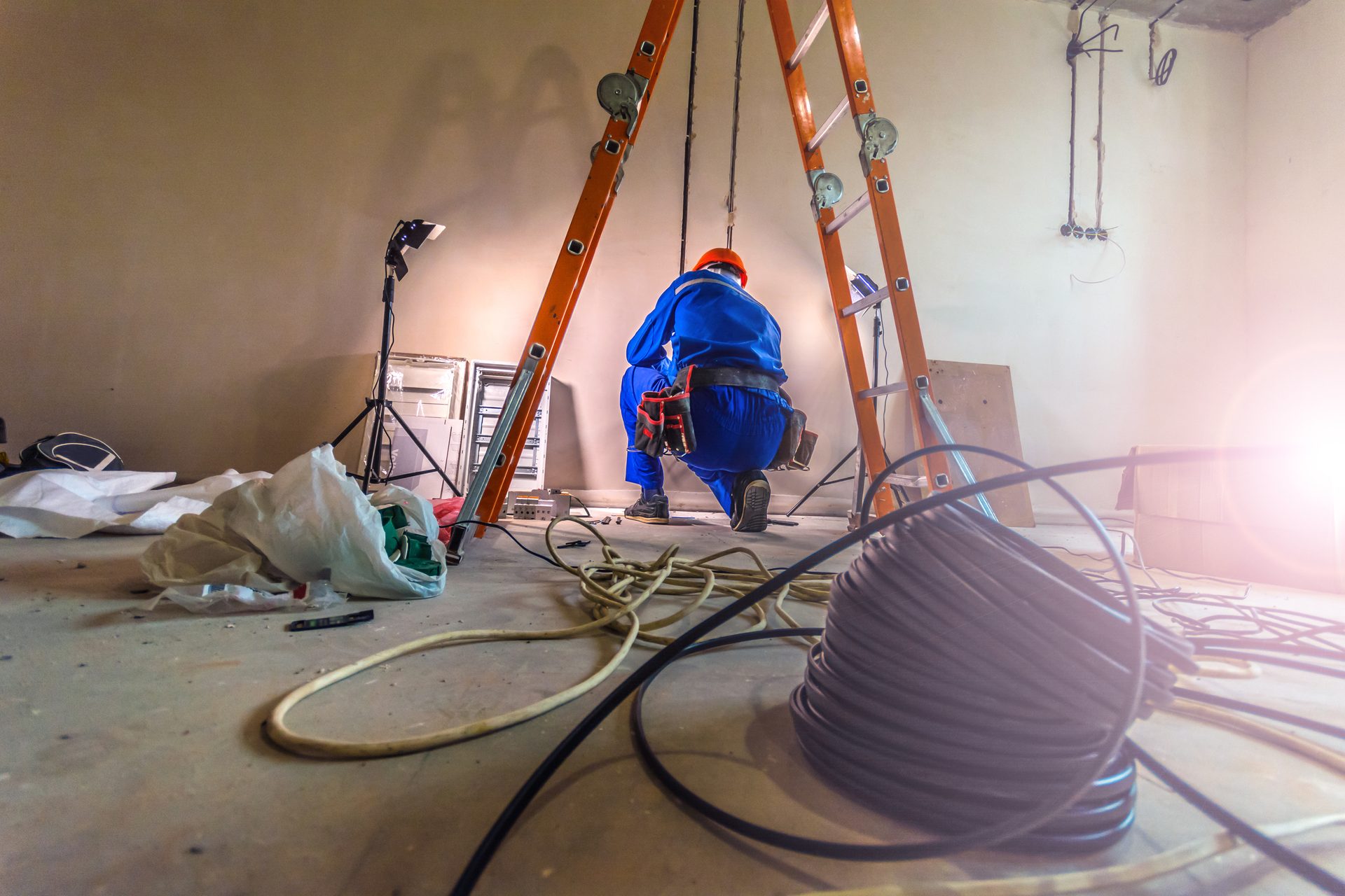 An electrician in blue overalls and an orange hard hat kneeling at a wall between two ladders, with cable reels on the floor of a room being renovated