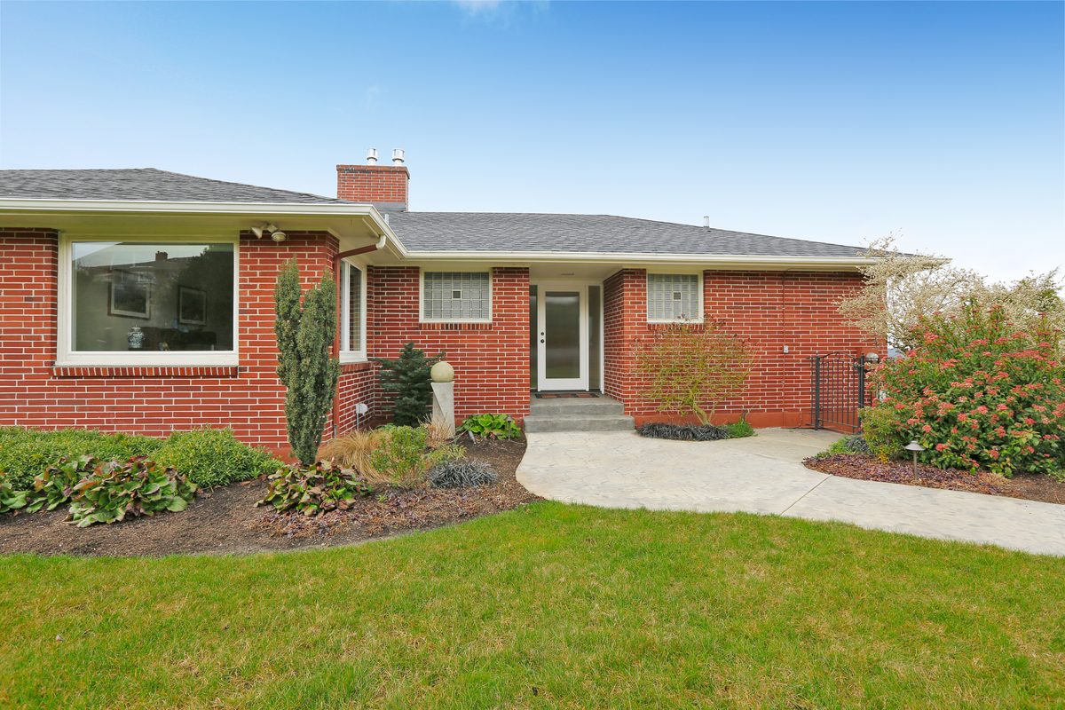 A single-storey red brick house with a low hipped roof, a chimney, a front lawn and garden beds along a curved path